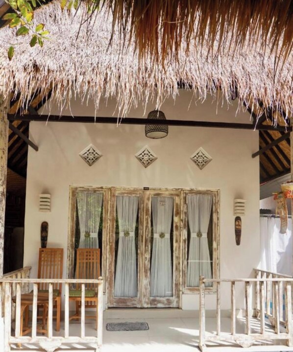 A covered outdoor area with a thatched roof, two doors with curtains, and a small seating area with wooden chairs. - Pondok Kana Homestay (Penida Island)