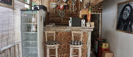 Une cuisine rustique avec un îlot central en bois, deux tabourets de bar, un réfrigérateur et un mur en bois orné d’un motif imitant des troncs d’arbres.