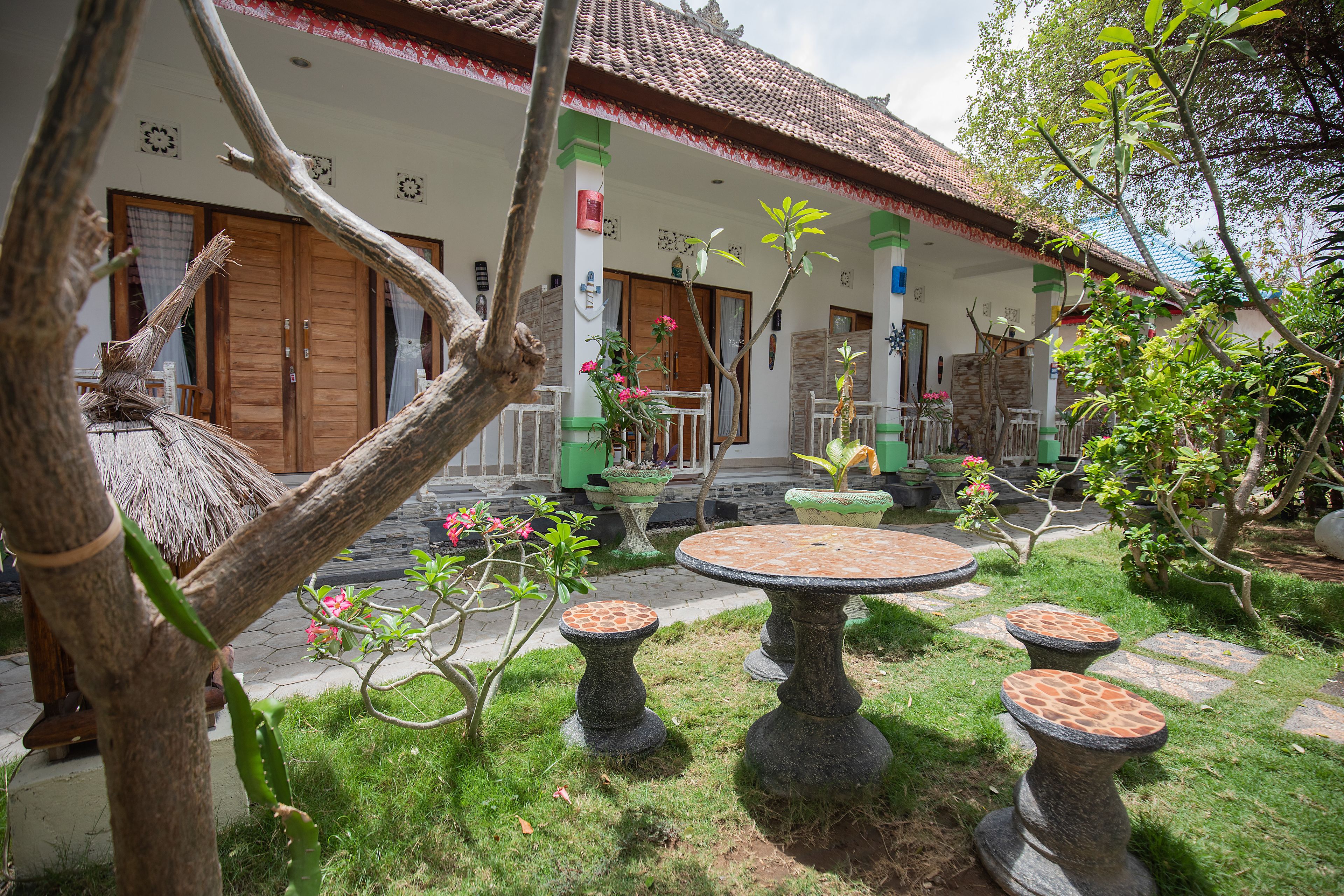 A courtyard with a thatched-roof structure, a stone table with four stools, and a variety of potted plants.