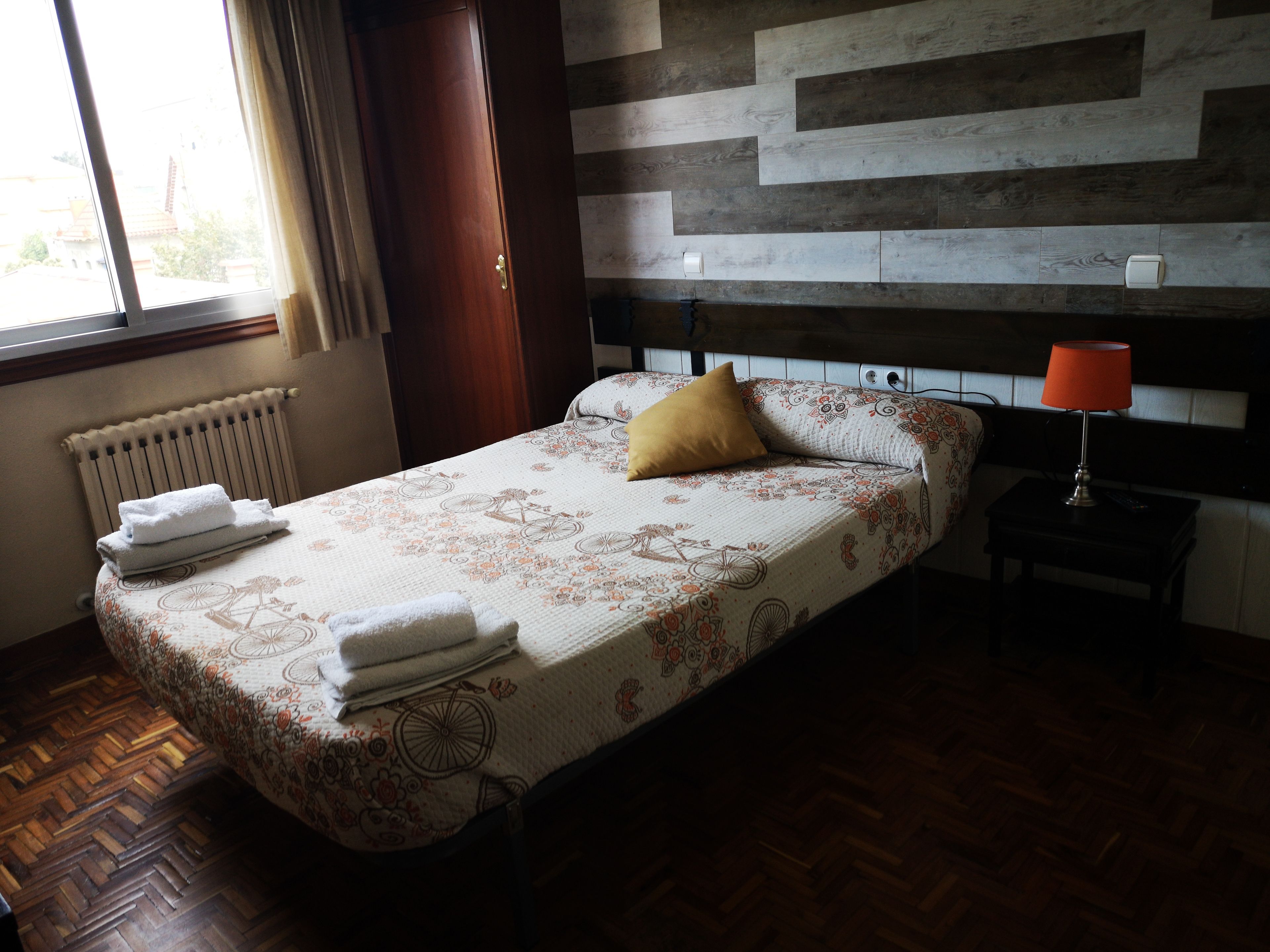 Una cama bien hecha con una colcha de estampado floral y una almohada amarilla. Se observa un cabecero de madera, una mesita de noche con una lámpara y un radiador en la pared.