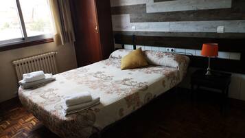 Una cama bien hecha con una colcha de estampado floral y una almohada amarilla. Se observa un cabecero de madera, una mesita de noche con una lámpara y un radiador en la pared.