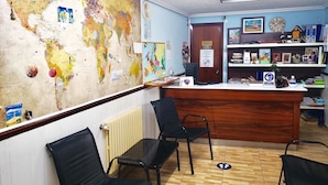 A reception area with a large world map, a wooden reception desk, black chairs, and a blue wall with shelves containing books and a clock. - Hostal Louro (O Porrino)