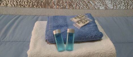 A neatly made bed with a blue towel, a white towel, and two bottles of shampoo.