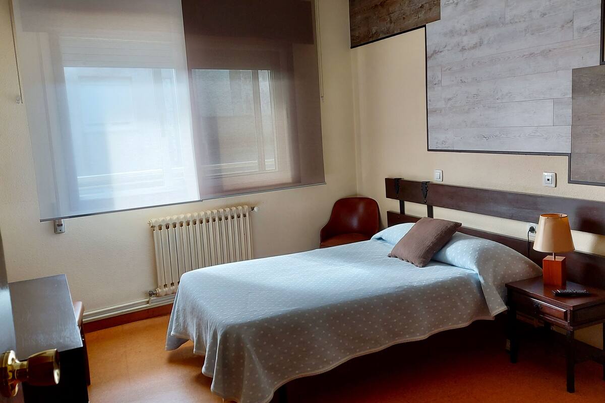A hotel room with a bed, a nightstand, a lamp, a chair, and a window with blinds.