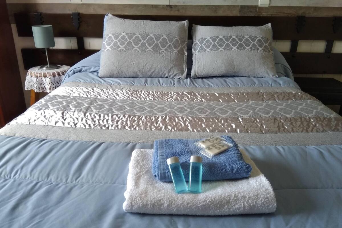 A neatly made bed with a patterned bedspread, two pillows, and a folded towel with toiletries on a bedside table.