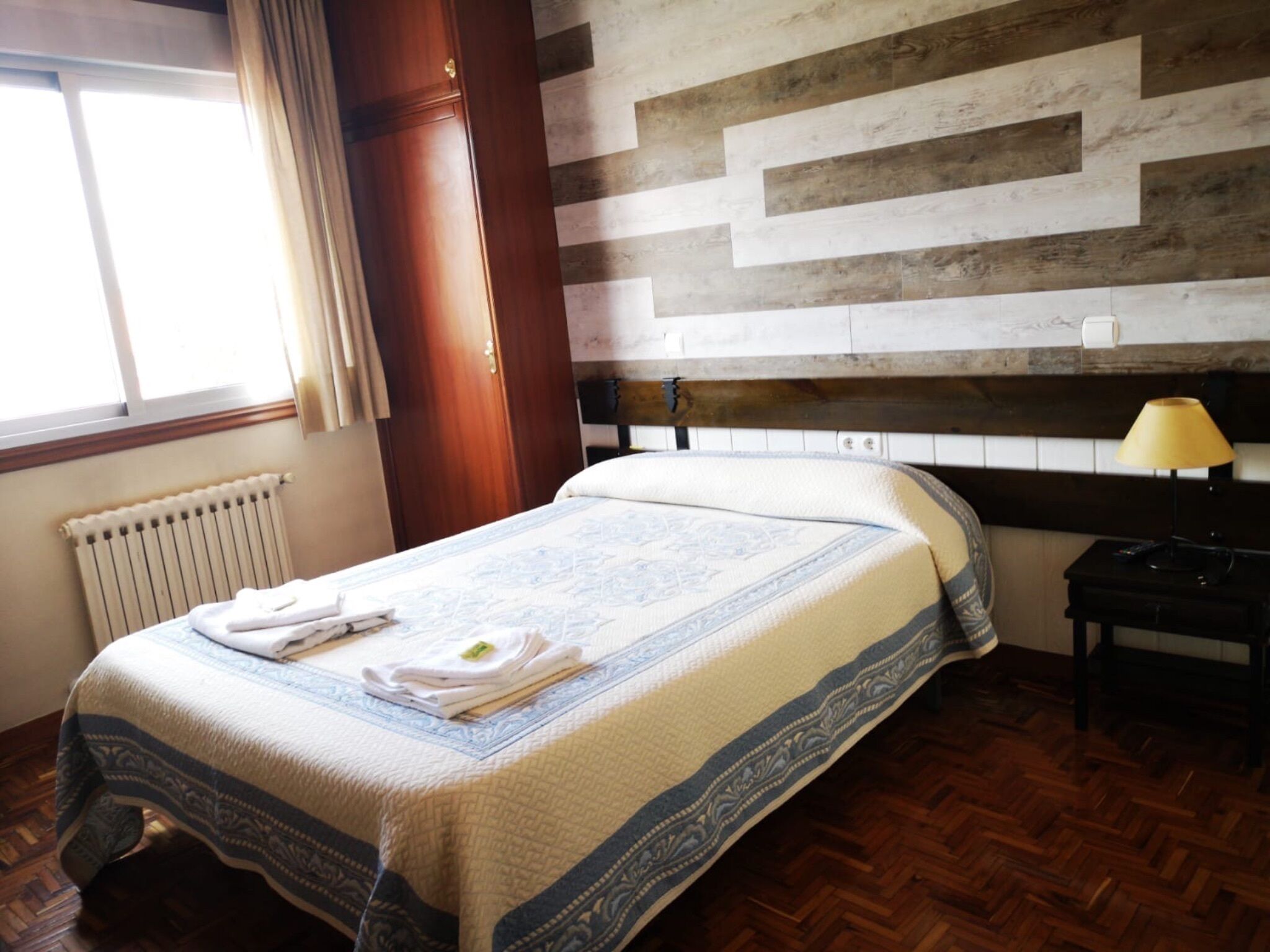 Una cama bien hecha con colcha estampada, un radiador, suelo de madera y una pared con patrón de espiga.