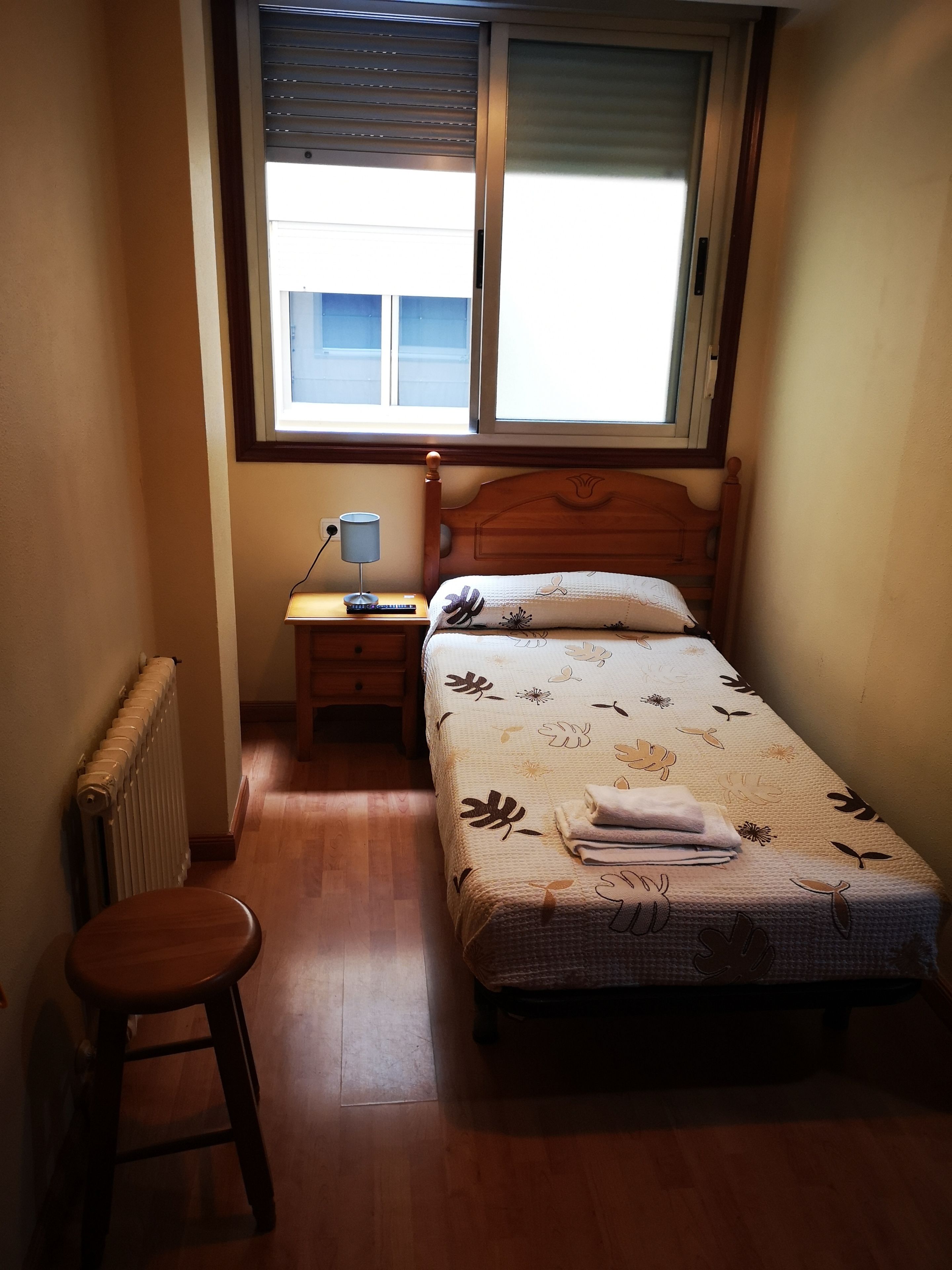 Un dormitorio pequeño con una cama individual, una mesita de noche, un escritorio con lámpara y un radiador.