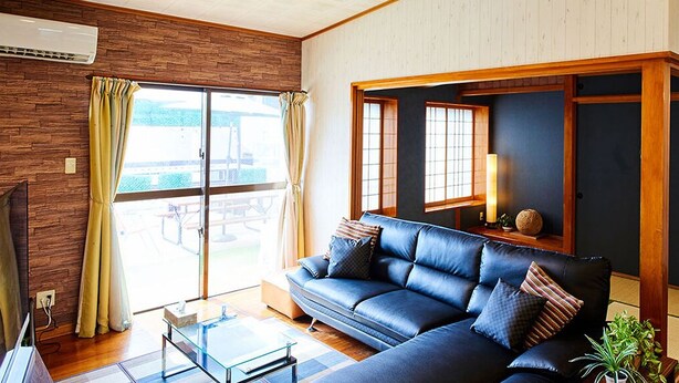 A living room with a dark blue sofa, a glass coffee table, and a TV. There is a sliding glass door leading to an outdoor area. - Terrace House Tsubokawa (Naha)
