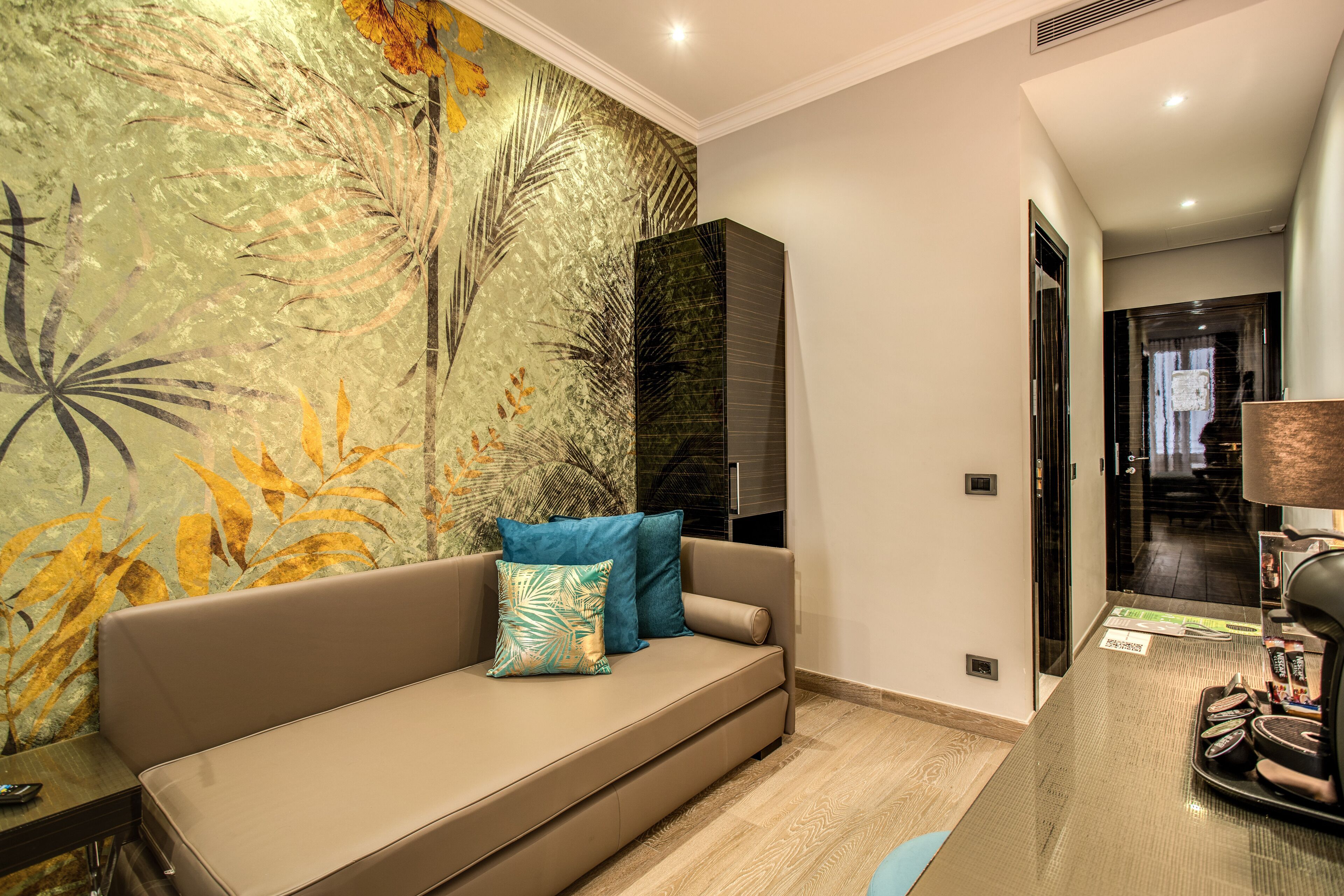 Una habitación de hotel moderna con sofá color beige, papel tapiz con estampado de hojas tropicales, pared de acento negra y una mesa de centro de vidrio con una cafetera.