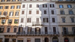 Um edifício de vários andares com entrada central, lojas no térreo e varandas nos andares superiores.