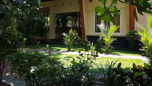 Ein gelbes Gebäude mit überdachter Terrasse, umgeben von üppiger Vegetation und Topfpflanzen.