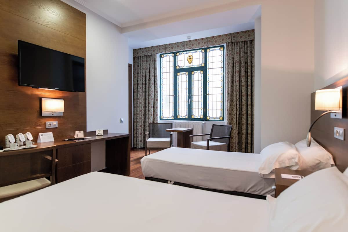 A hotel room with two beds, a desk, a TV, and a window with curtains.
