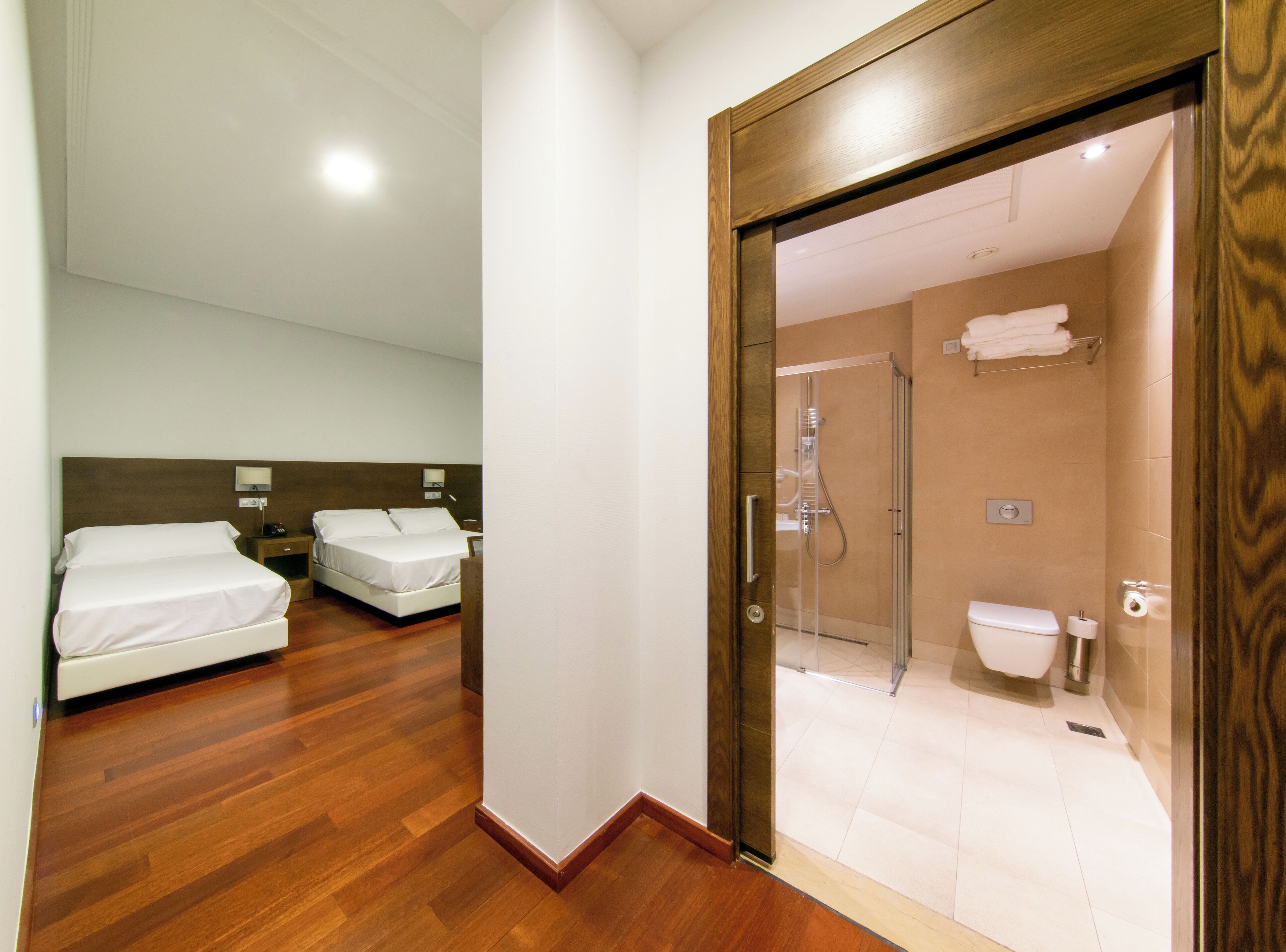 Ein Hotelzimmer mit zwei Betten, Holzboden und einer Glastür zu einem Badezimmer mit Dusche und Toilette.