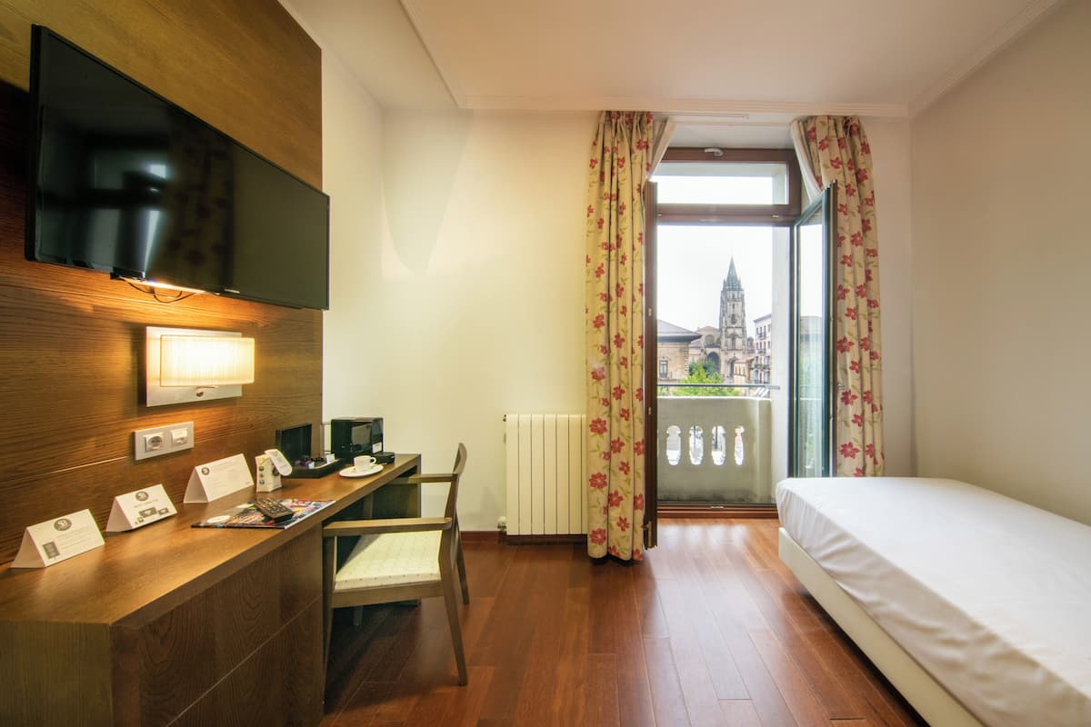 A hotel room with a wooden desk, a bed, a television, and a view of a building through the window.
