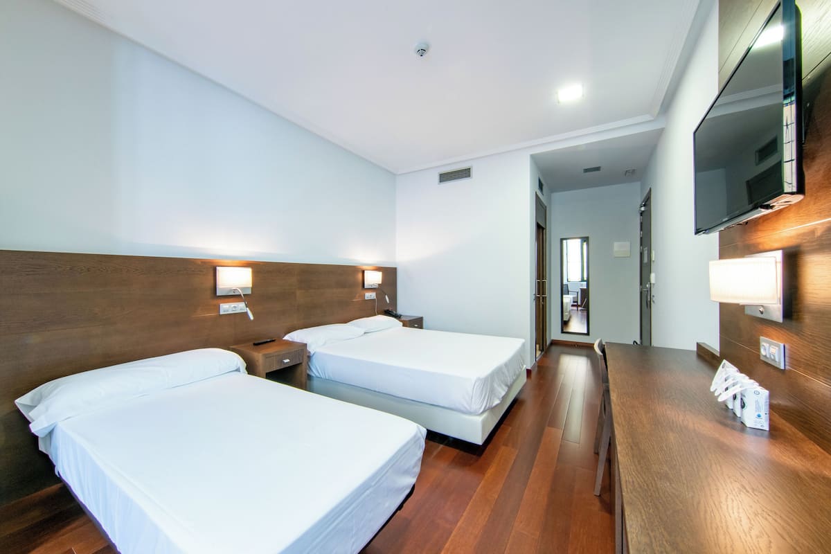 A hotel room with two beds, wooden flooring, a wooden headboard, a TV mounted on the wall, and a desk with a lamp.