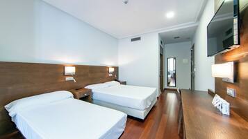 A hotel room with two beds, wooden flooring, a wooden headboard, a TV mounted on the wall, and a desk with a lamp.