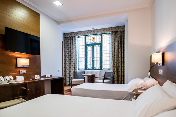 Habitación de hotel con dos camas, un escritorio, un televisor de pantalla plana, una mesita y una ventana con cortinas.
