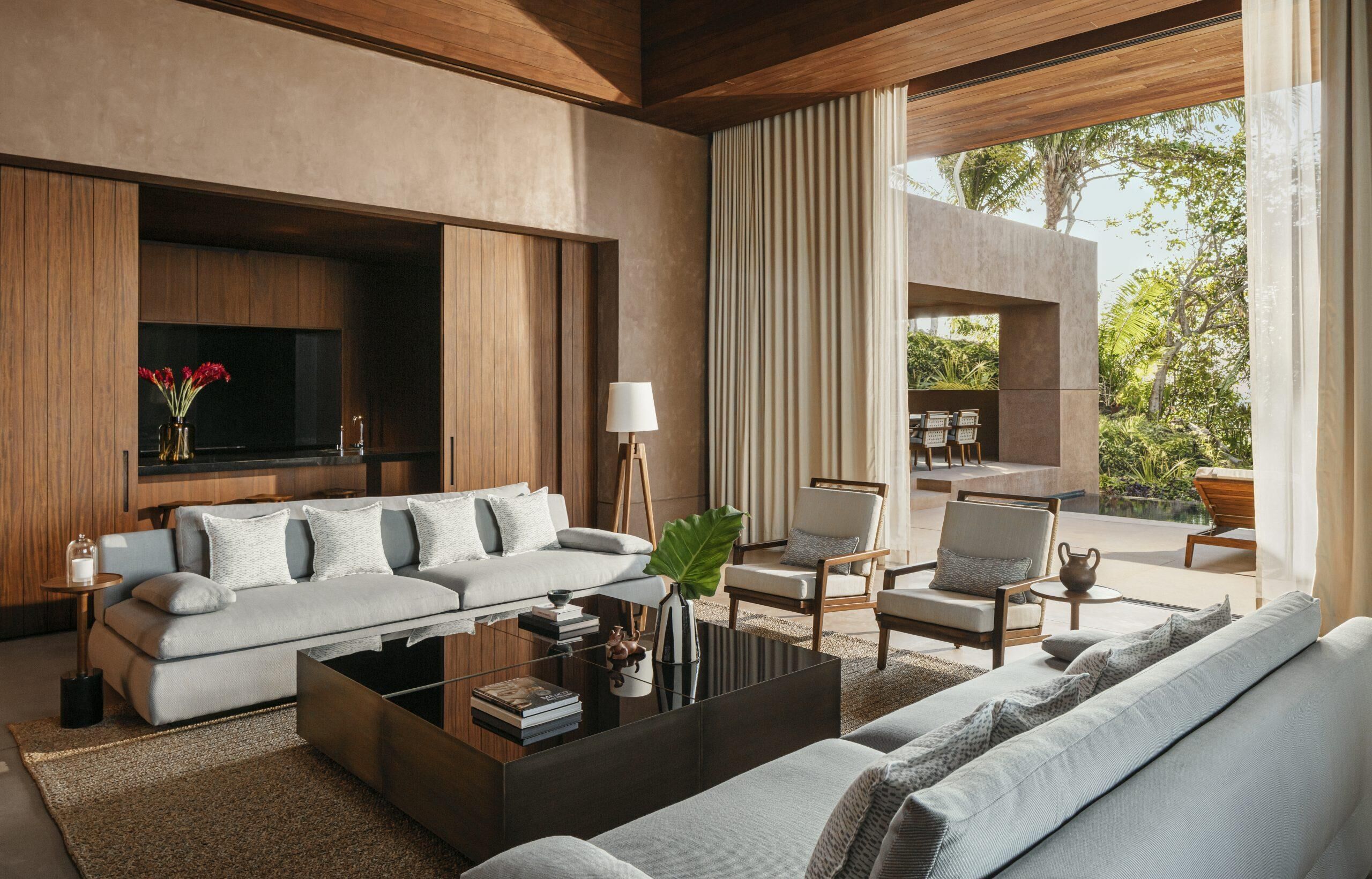 A modern living room with a sofa, coffee table, and chairs. There is a television mounted on the wall and a window with curtains.