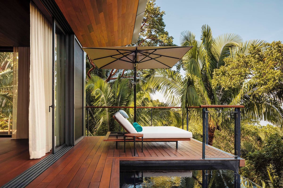 Un balcón con cubierta de madera, una silla de descanso, un gran paraguas y vistas a exuberante vegetación y una piscina.