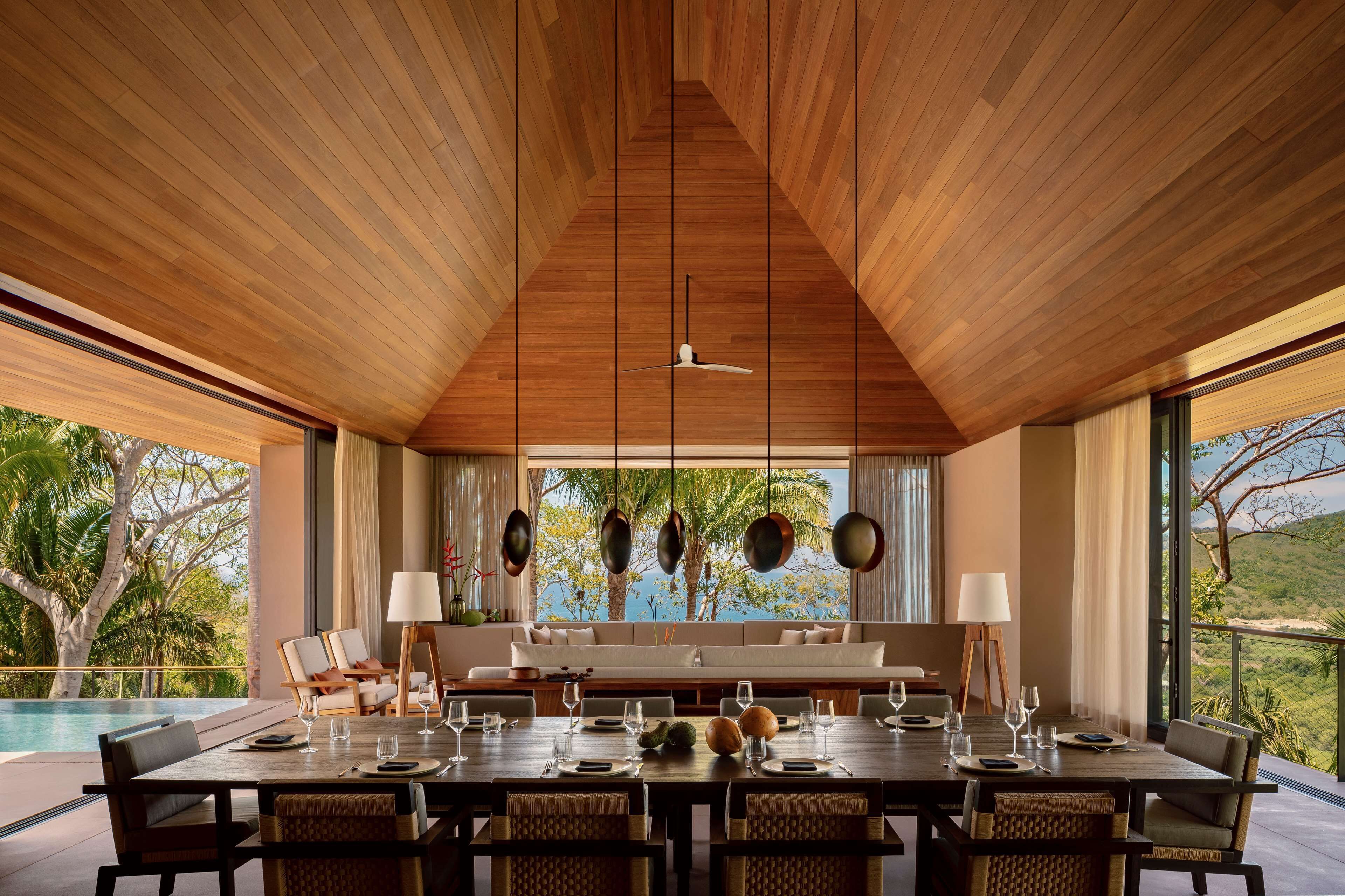 A dining area with a high ceiling, wooden beams, and a view of palm trees and a pool.