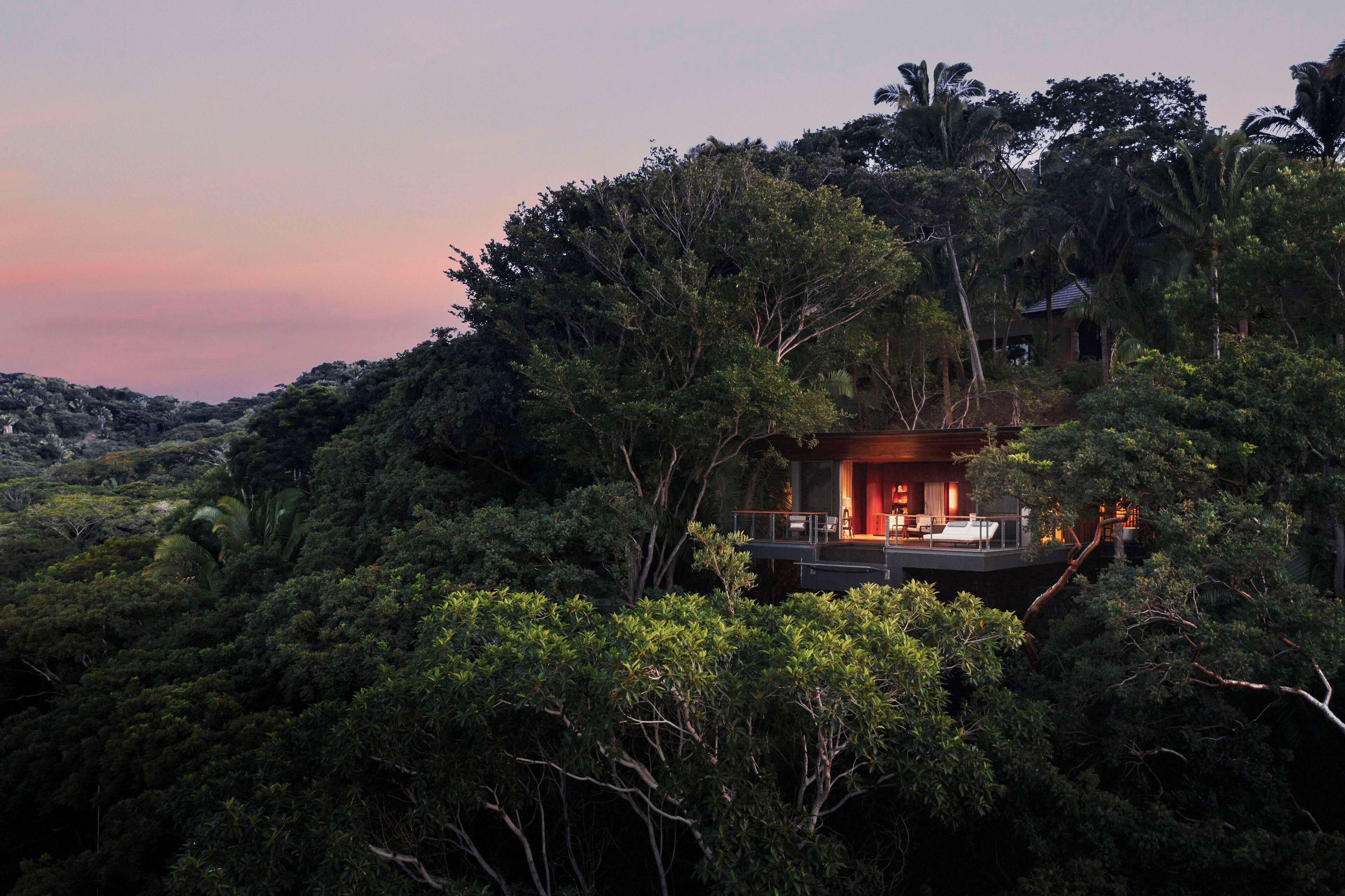 A secluded, modern house surrounded by dense forest, featuring a large balcony with a seating area and a visible interior with warm lighting.