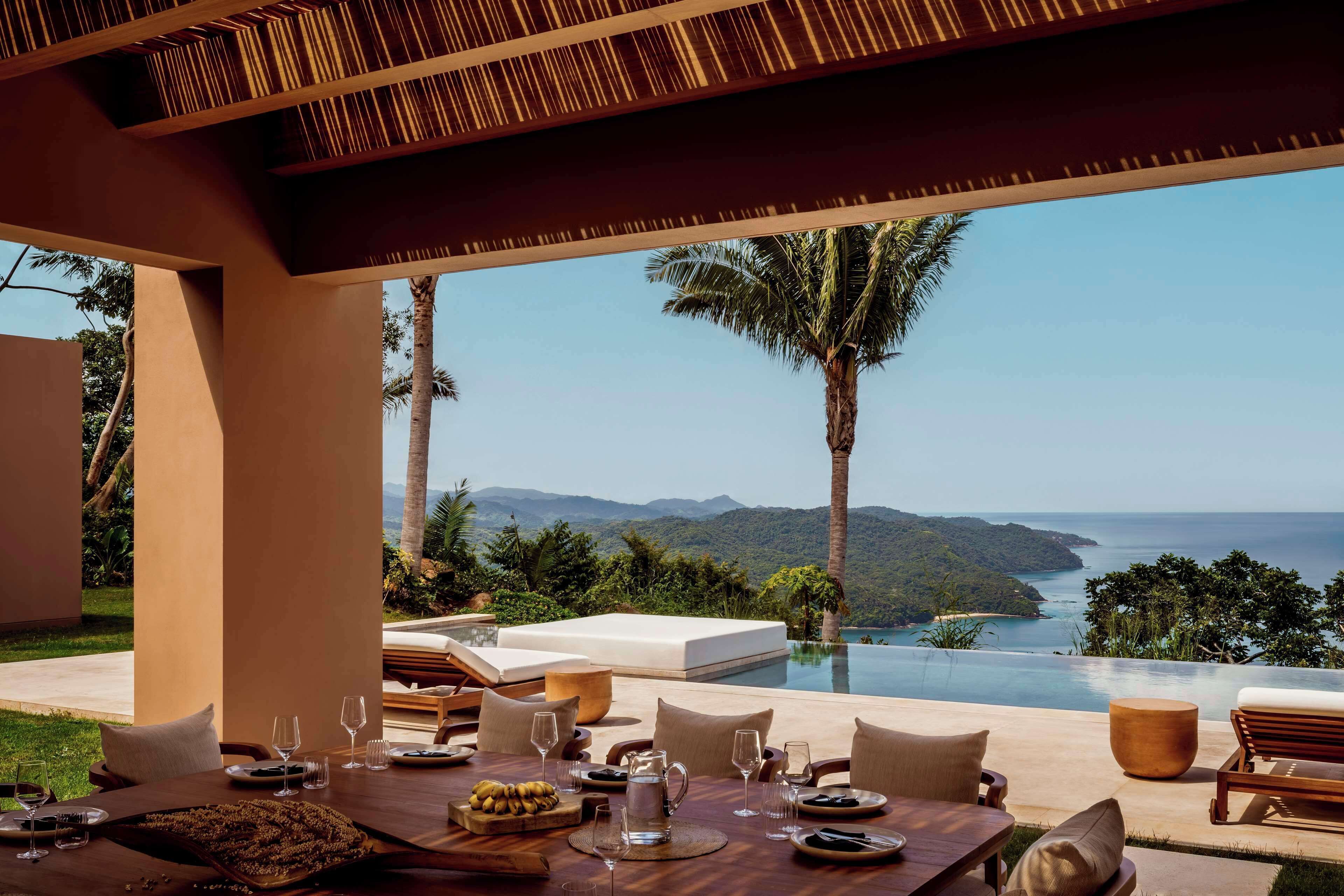 A dining area with a table set for a meal, overlooking a pool with a view of the sea and mountains.