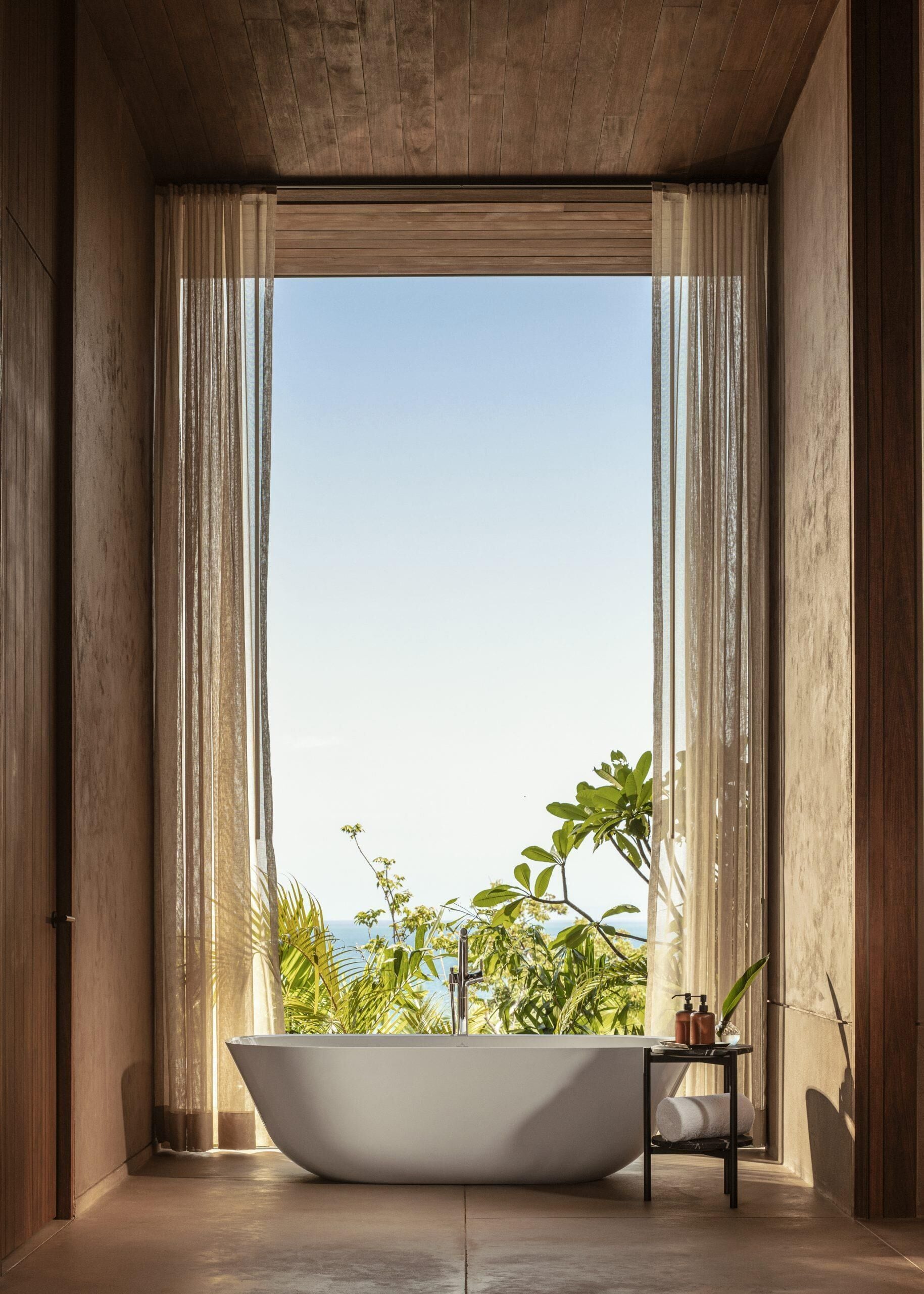 Un baño moderno con una bañera independiente, una mesita con artículos de aseo y vistas a la vegetación y al cielo a través de grandes ventanales.