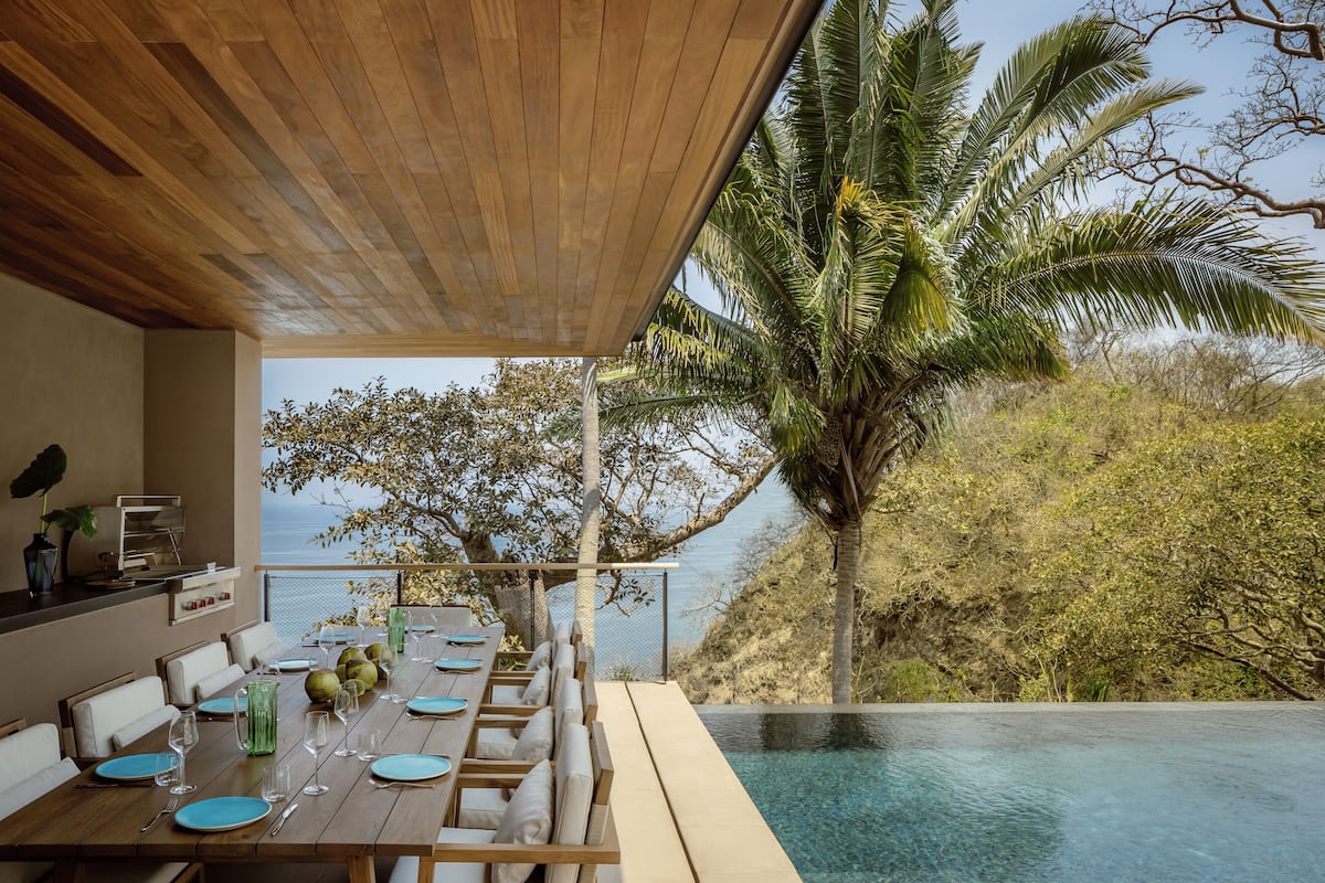 Un área de comedor al aire libre con una mesa de madera preparada para seis personas, con vistas a una piscina y un paisaje pintoresco del mar y palmeras.