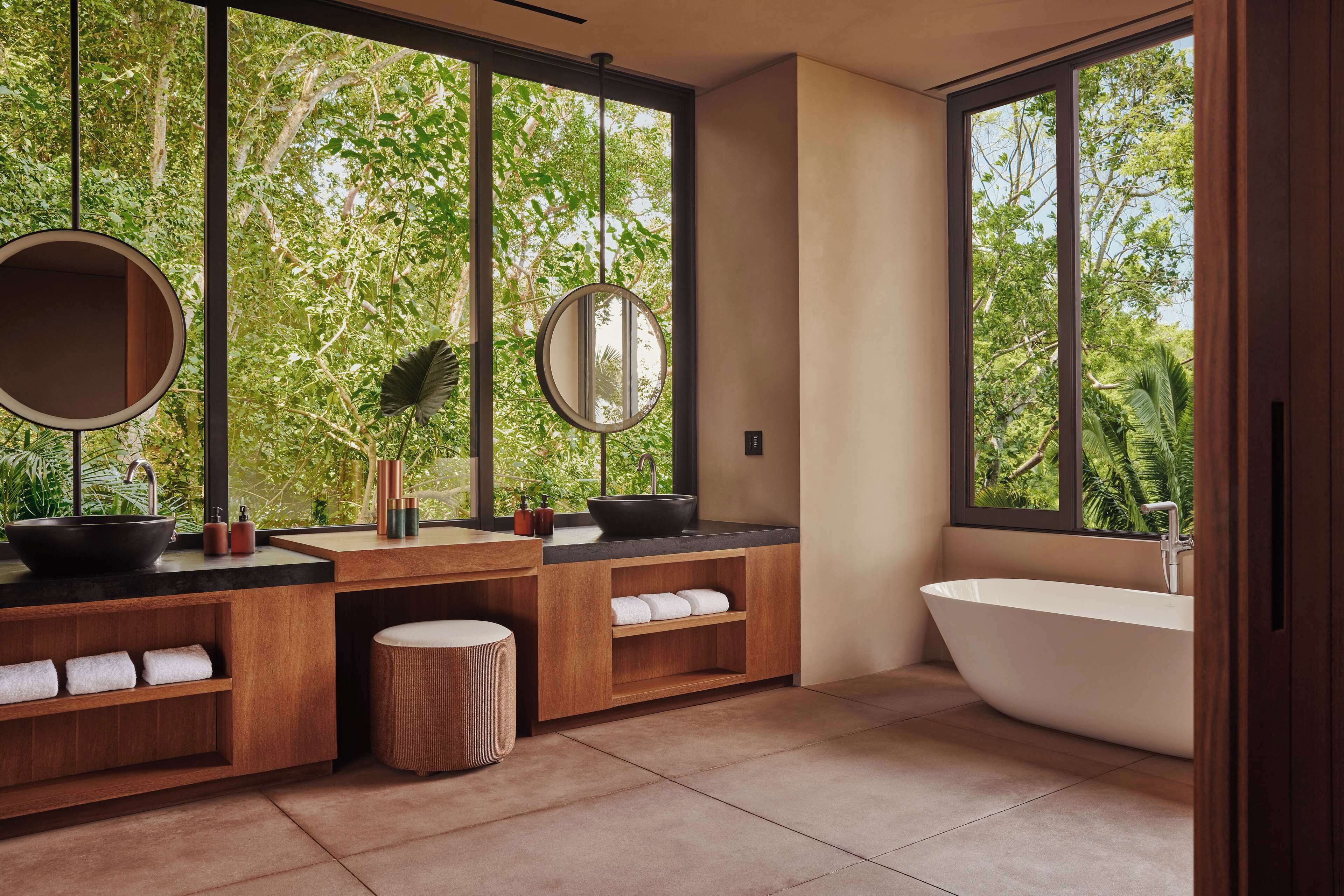 A modern bathroom with a freestanding bathtub, wooden vanity with a round mirror, and a large window offering a view of greenery.
