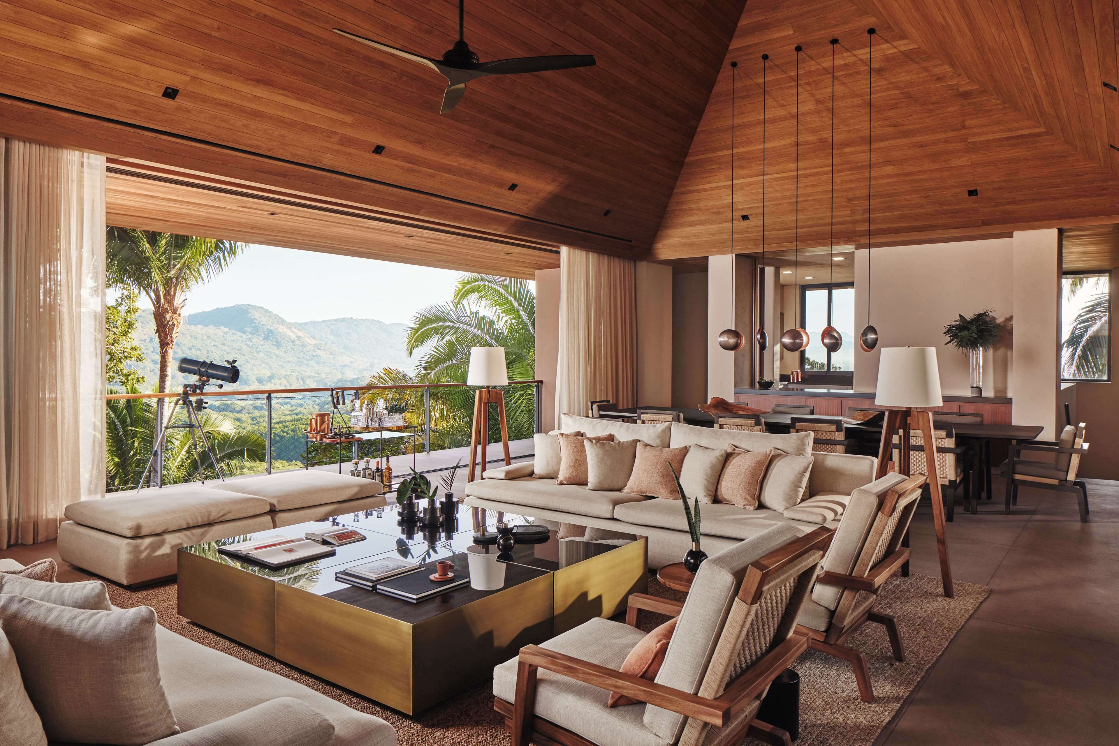 A spacious living area with a large sofa, coffee table, and dining set. A balcony with a view of mountains and palm trees is visible.