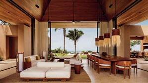 A spacious living area with a large wooden ceiling, a dining area with wooden furniture, and a view of palm trees and the ocean. - One&Only Mandarina (Lo de Marcos)