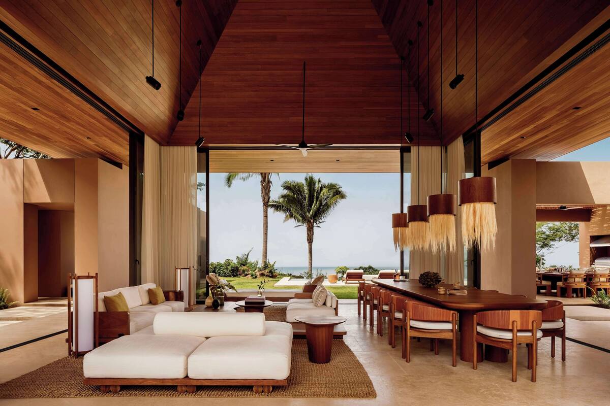A spacious living area with a large wooden ceiling, a dining area with wooden furniture, and a view of palm trees and the ocean.