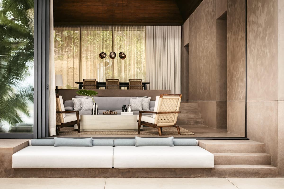 Una sala moderna con sofá, sillones y una mesa de centro. Hay una zona de comedor con sillas y una mesa. La habitación tiene ventanas amplias y vistas al exterior con vegetación.