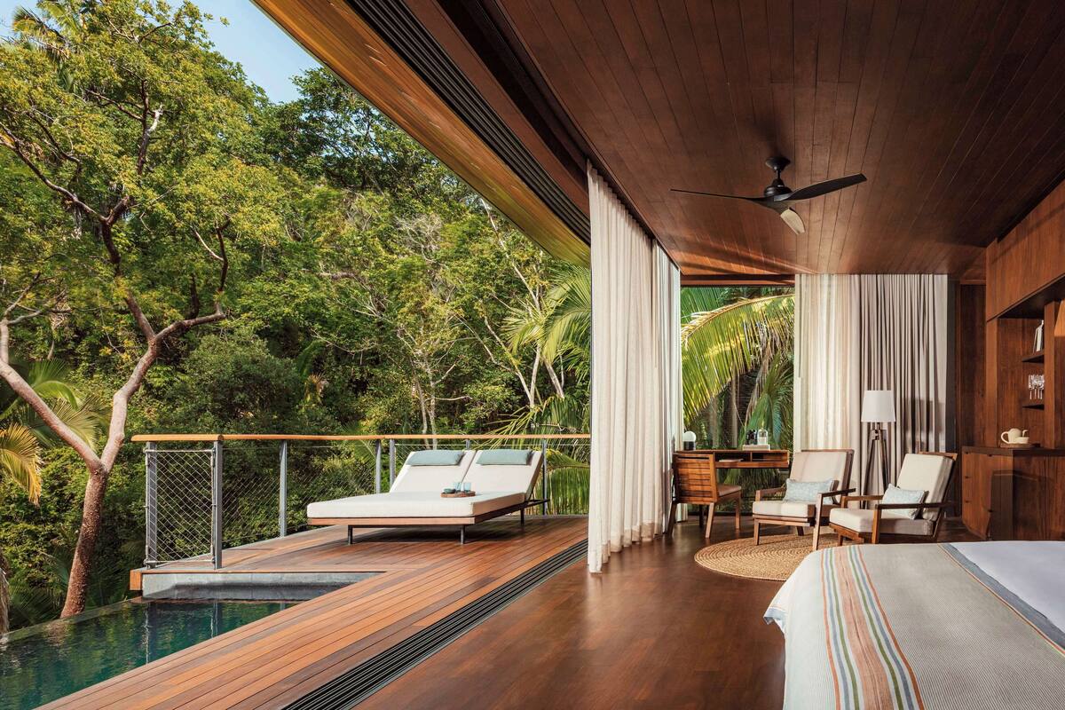 A bedroom with a large bed, a wooden ceiling, and a view of a pool and trees.