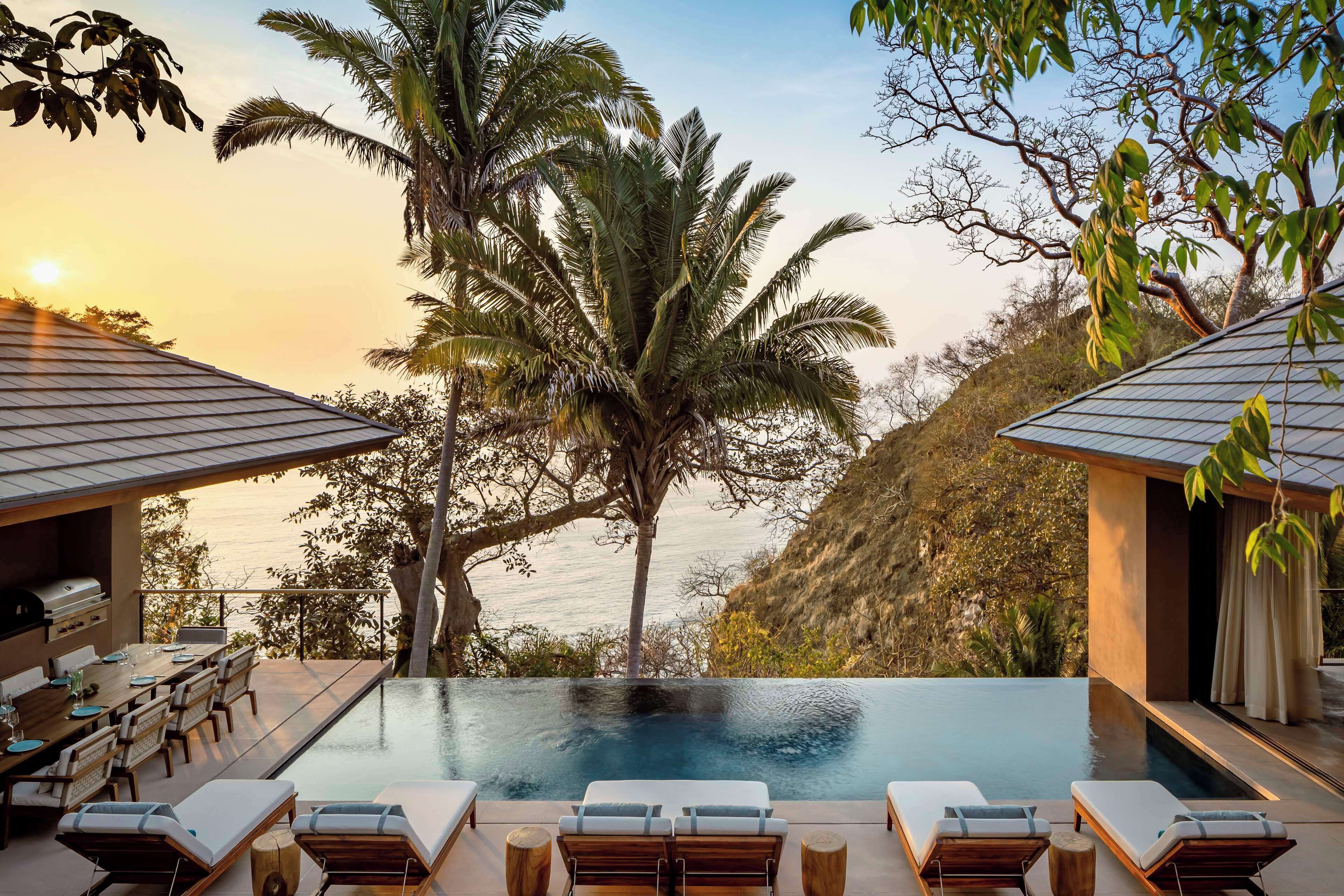 Una piscina infinita con sillones de descanso, un área de comedor con parrilla y vistas a palmeras y al océano.