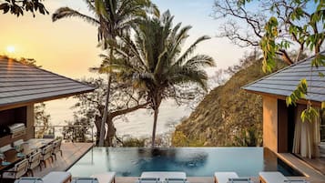 Una piscina infinita con sillones de descanso, un área de comedor con parrilla y vistas a palmeras y al océano.