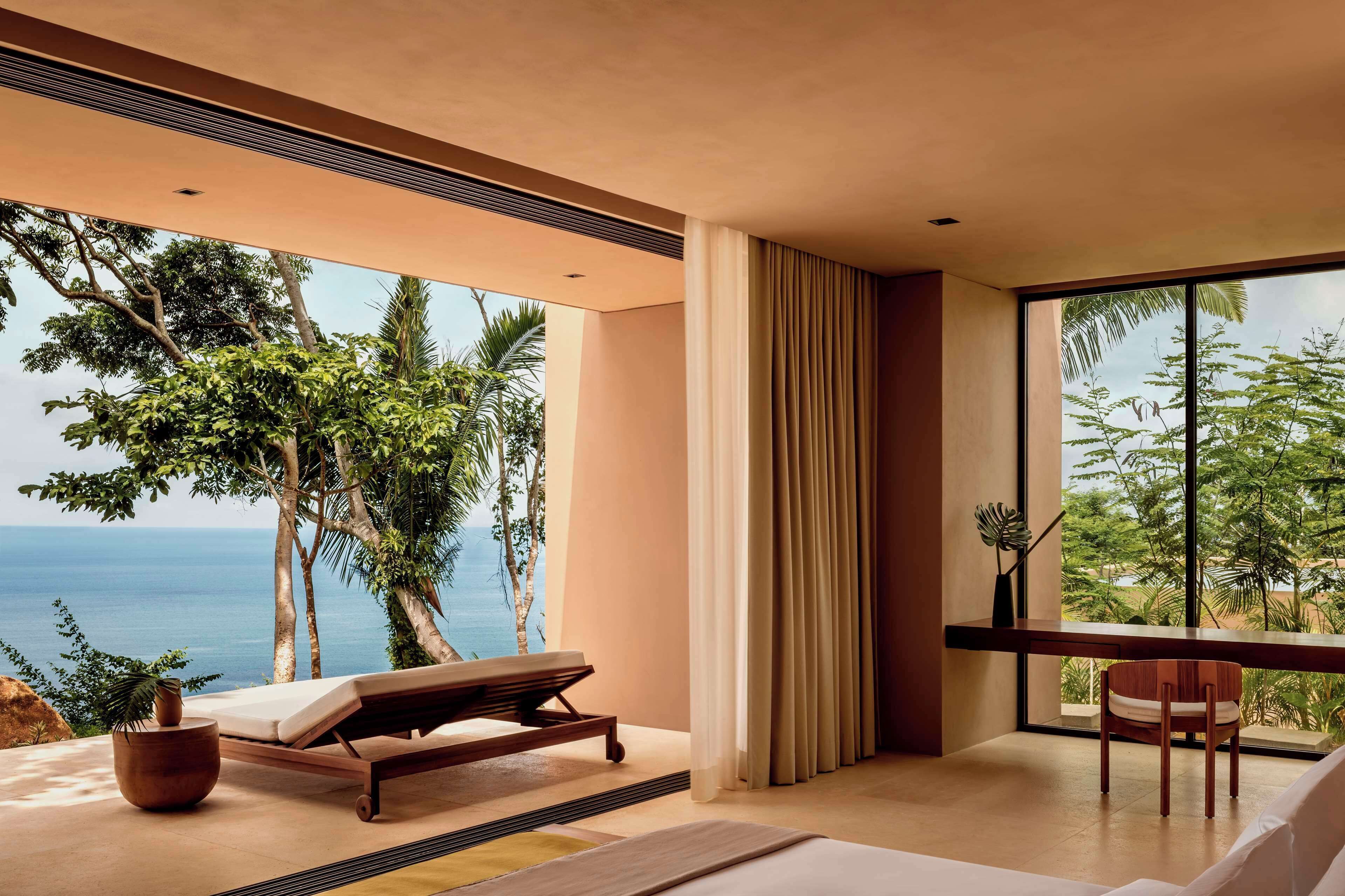 Quarto moderno com uma grande janela com vista para o mar, uma cama, uma poltrona e uma escrivaninha de madeira.
