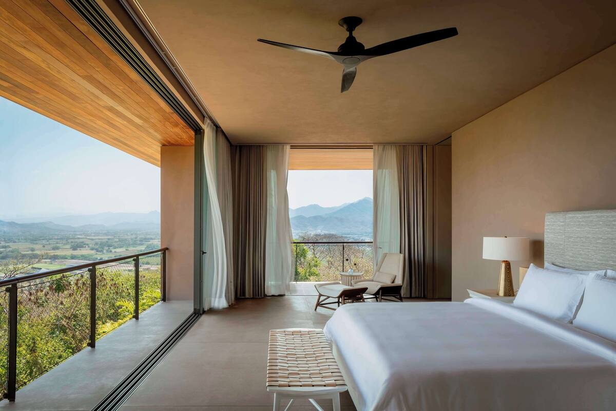 A hotel room with a large bed, a ceiling fan, a balcony with a view of the mountains, and a table with chairs.