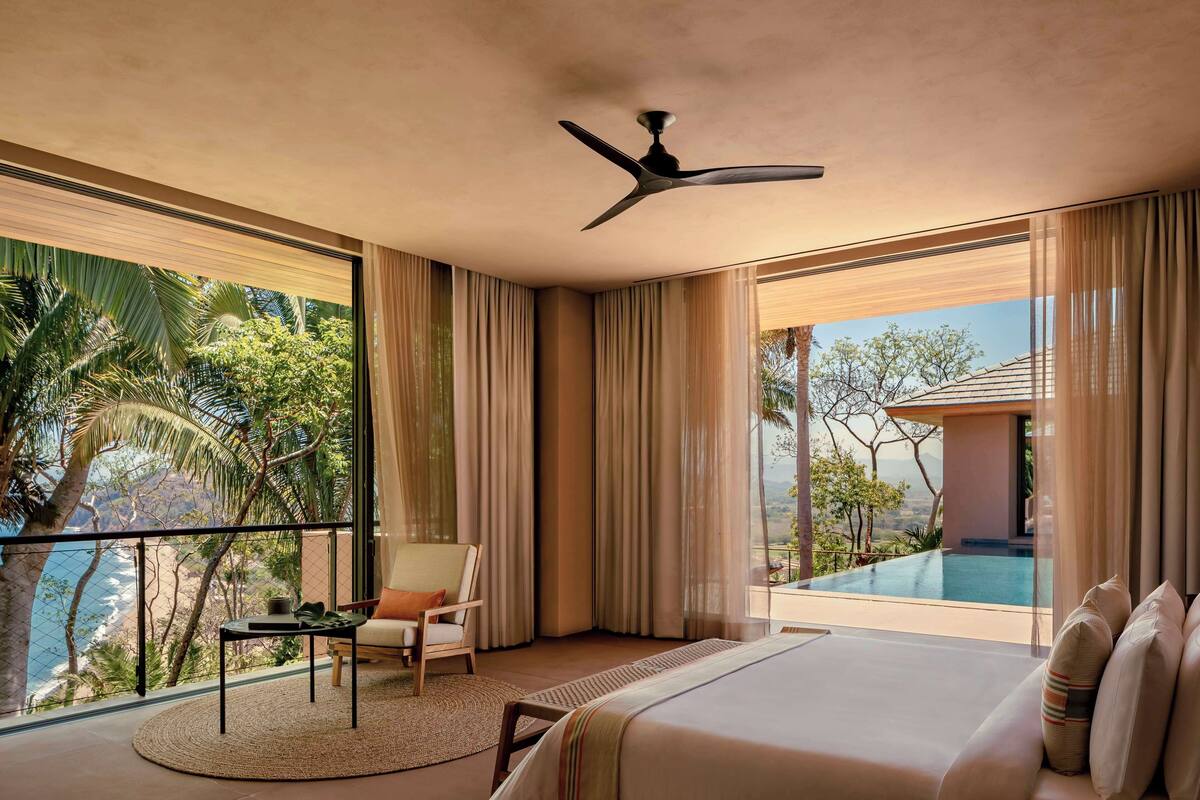 A modern hotel room with a large bed, a ceiling fan, a balcony with a view of the ocean, and a small table with a chair.