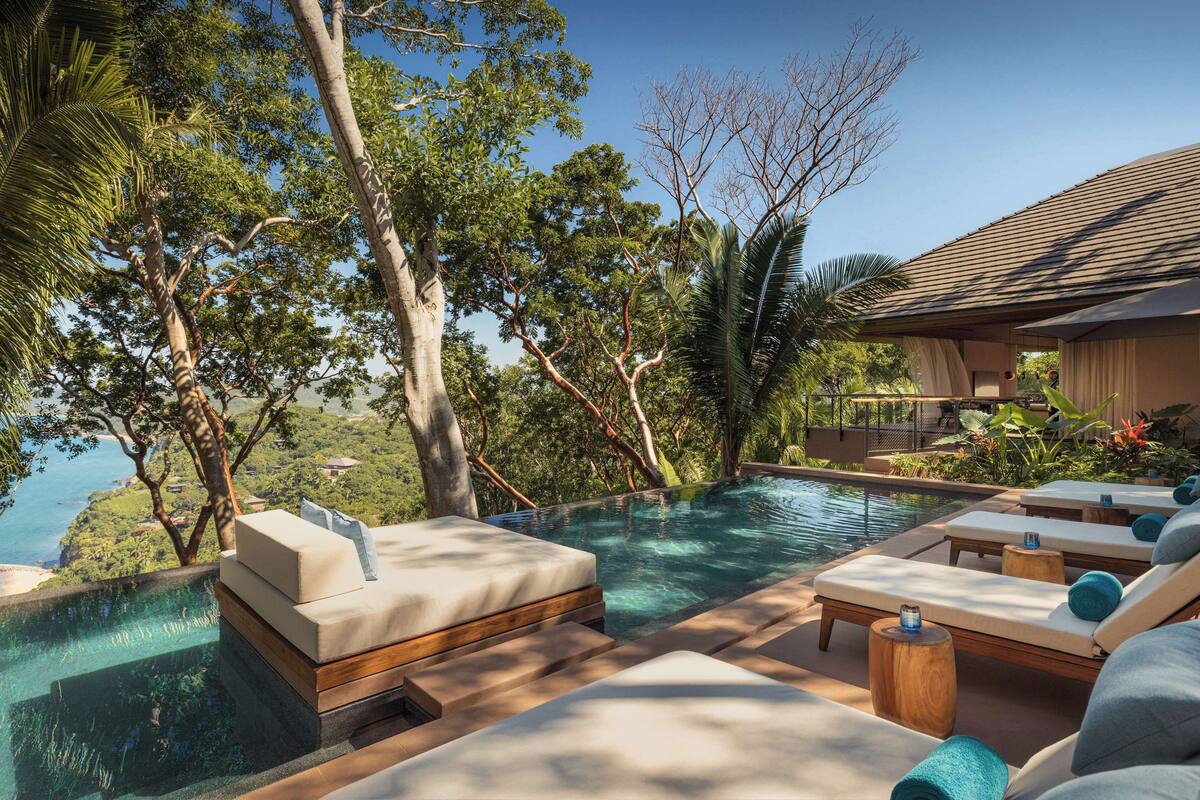 An infinity pool with lounge chairs, surrounded by tropical trees and a view of the landscape.