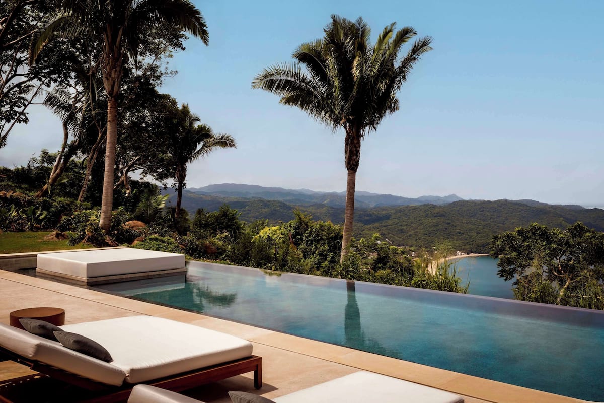 Piscina infinita con sillas de descanso, rodeada de palmeras y vistas a un lago y montañas.