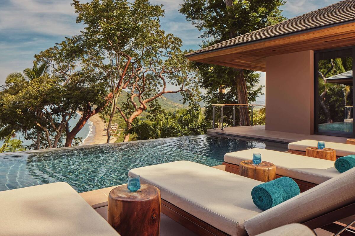 A poolside area with lounge chairs and wooden stools, overlooking a beach and lush trees.