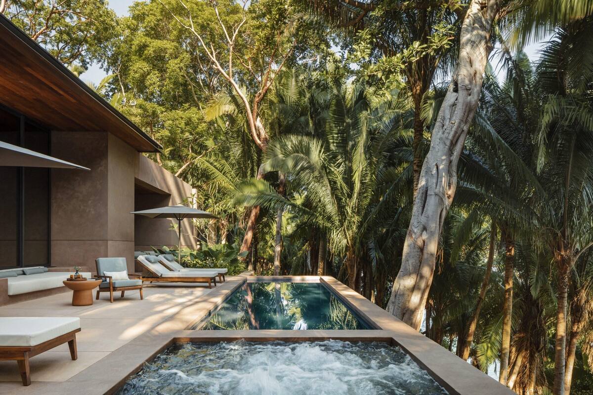 A modern outdoor pool area with lounge chairs and a hot tub, surrounded by lush tropical trees.