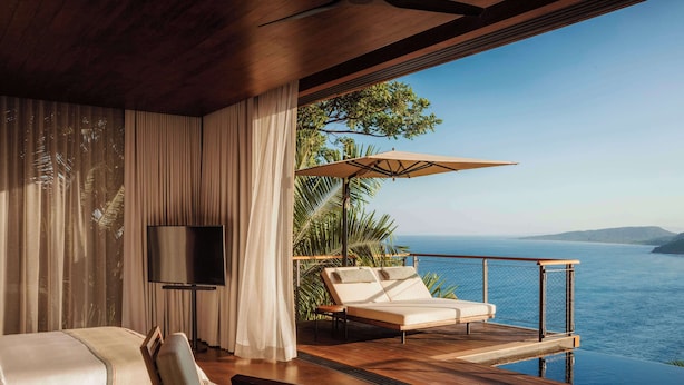 Una habitación lujosa con una cama grande, televisión, terraza de madera, sillón y vistas al mar.