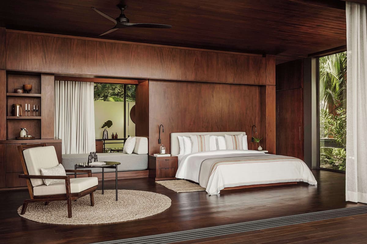 Un espacioso dormitorio con una cama grande, una zona de estar con una silla y una mesa, paneles de madera y vistas por una ventana amplia.