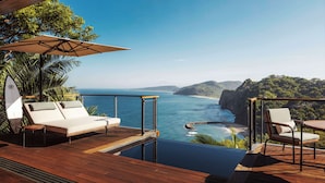 Terraza de madera con un sofá blanco y una mesa, con vistas a la costa, acantilados y una playa.