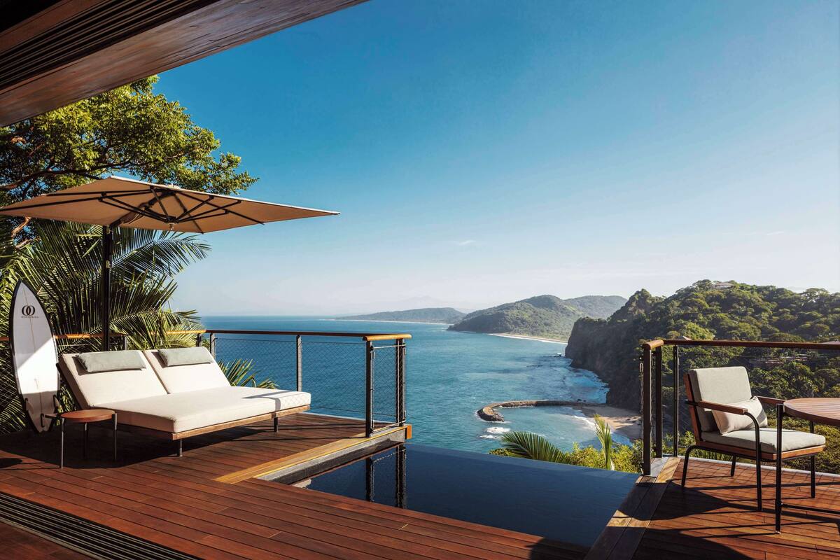 A wooden deck with a white sofa and a table, overlooking a coastal view with cliffs and a beach.