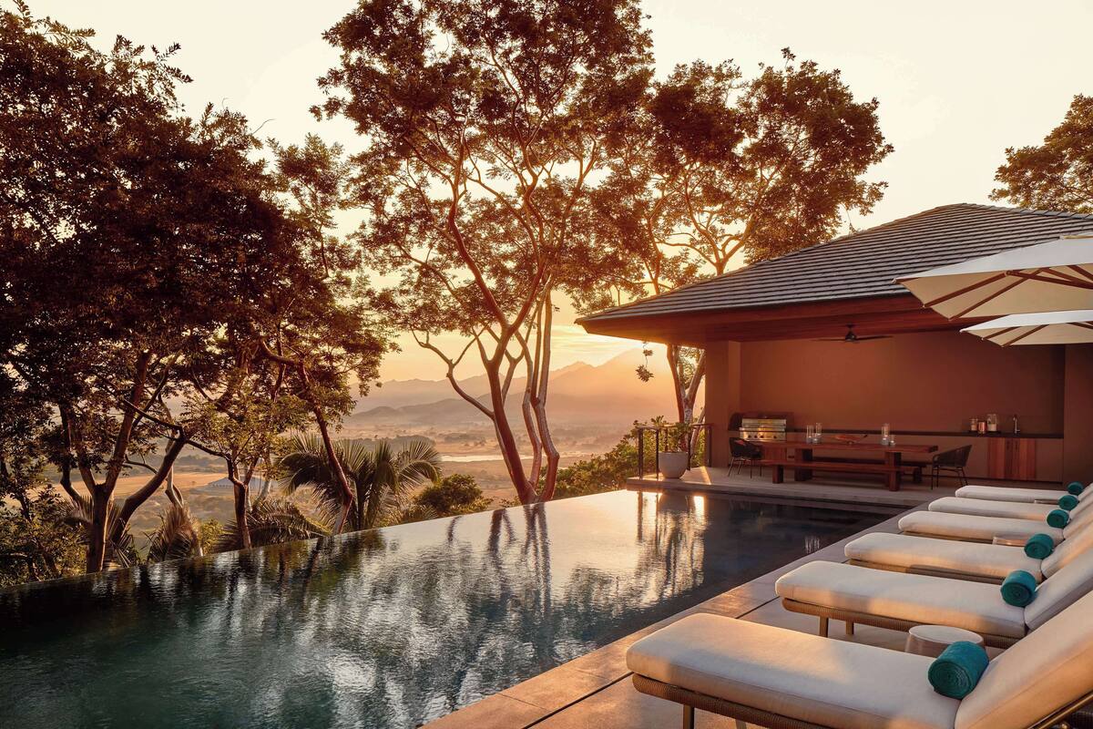 An infinity pool with lounge chairs, a dining area, and a view of trees and mountains.