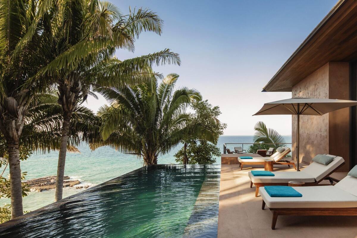 A poolside area with lounge chairs and an umbrella, overlooking a clear blue sea and palm trees.