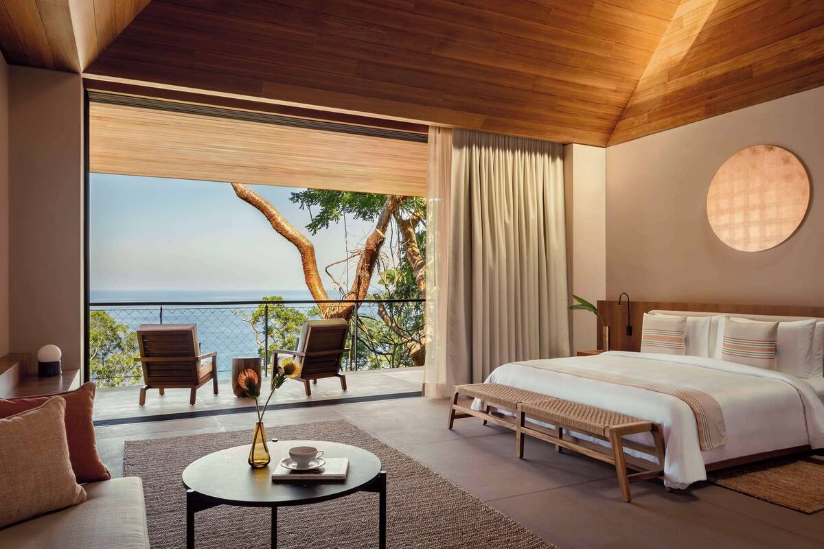 A modern bedroom with a large bed, a view of the sea, and a balcony with chairs.